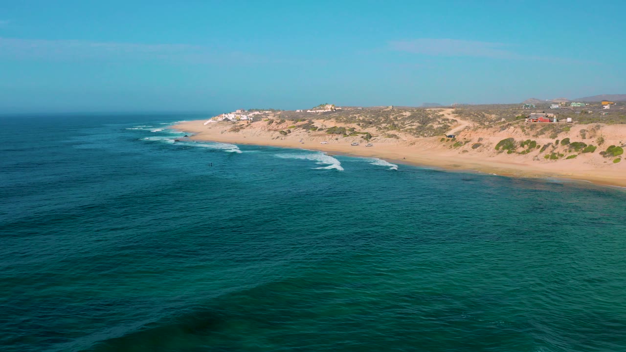 A wide aerial frame of Shipwrecks Beach with turquoise and deep blue water, thin surf foam near shore, pale sand, low dune vegetation, and no visible people.