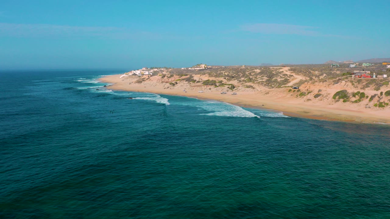 Opening aerial frame over Shipwrecks Beach in Baja California Sur, Mexico, with teal water in front, a long sandy shore, low dunes, distant buildings, and no visible people.