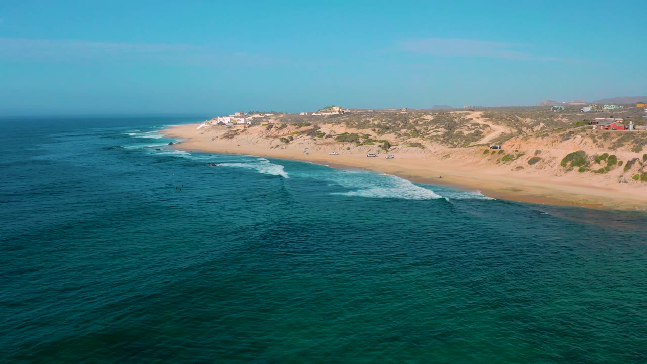 High aerial frame at Shipwrecks Beach showing deeper teal water in the foreground, foam bands near shore, tan dunes, small distant buildings, and no visible people.