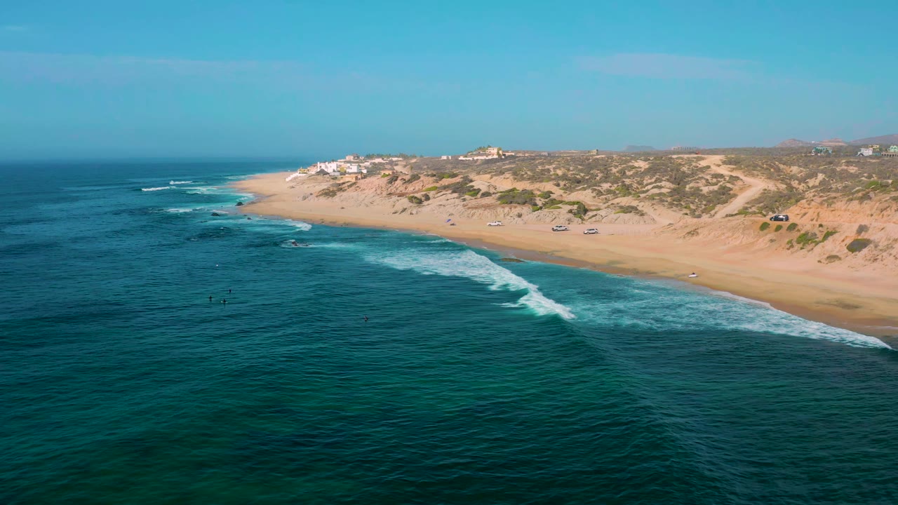 Drone view along a sandy shoreline with breaking waves and low dunes.
