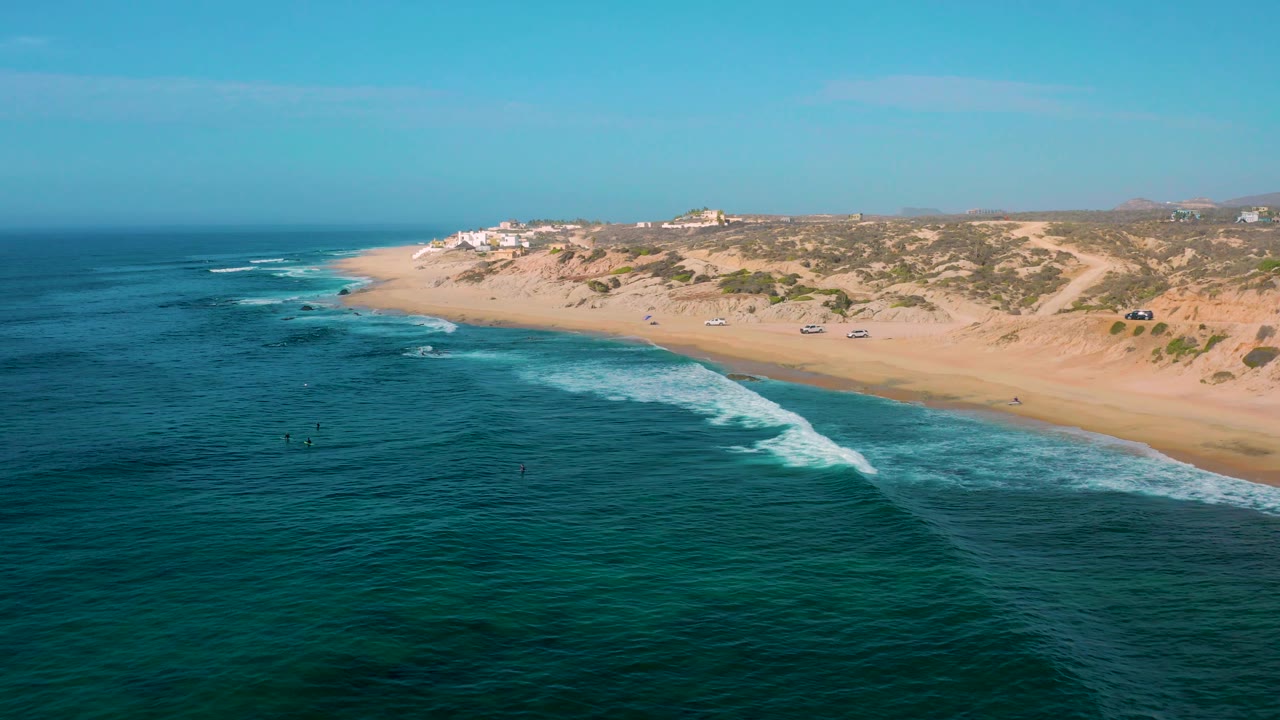 Wide aerial view of Shipwrecks Beach with darker ocean texture in the foreground, a white wave curling toward shore, tan sand, dune vegetation, and no visible people.