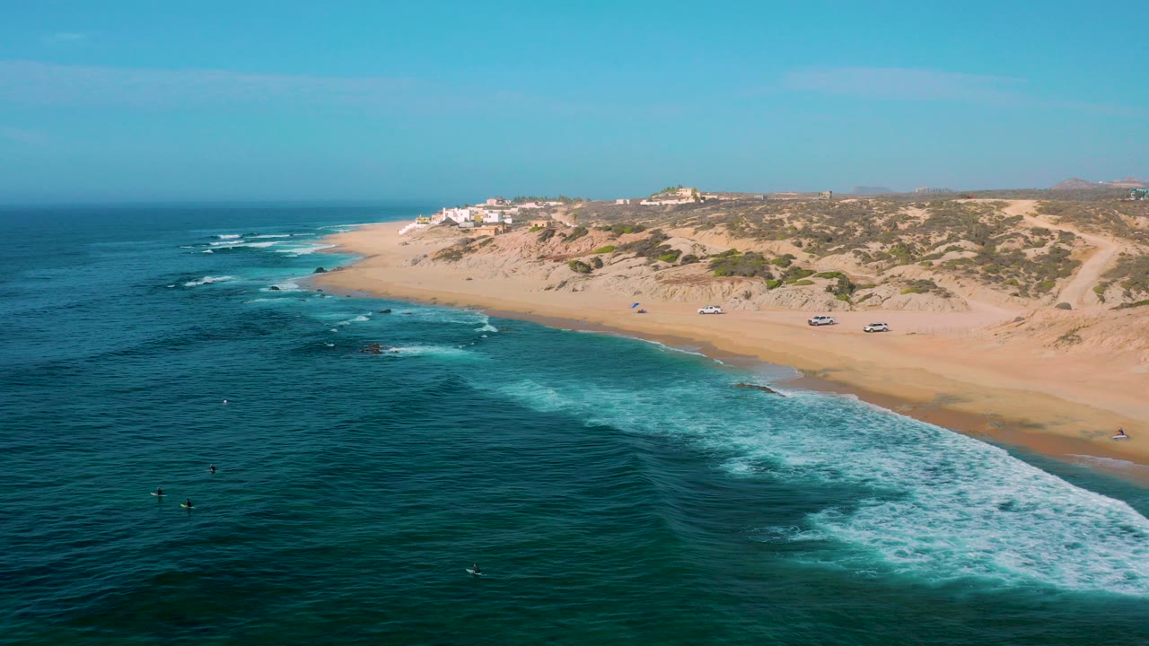 Opening aerial frame over Shipwrecks Beach in Baja California Sur, with blue ocean in front, white surf near rocks, a broad sandy beach, scrub dunes, distant ridge buildings, and no visible people.