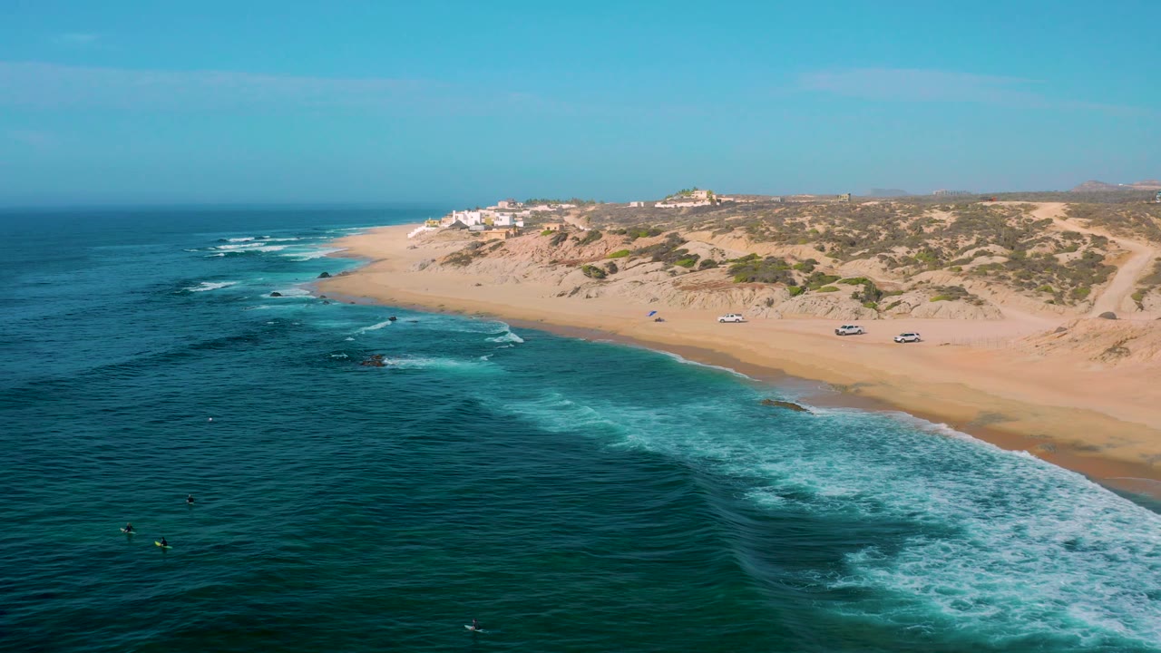 Wide aerial frame over Shipwrecks Beach with the surf line sweeping across the lower right, darker water offshore, tan dunes behind the beach, ridge-top buildings, and no visible people.
