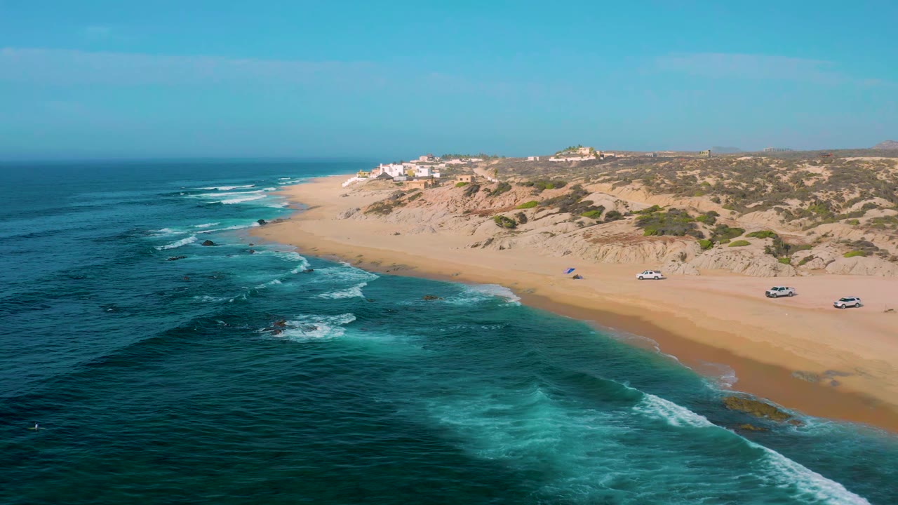 Aerial view at Shipwrecks Beach showing foamy surf bands moving toward a broad beach, with dune ridges, small parked vehicles, distant buildings, and no visible people.