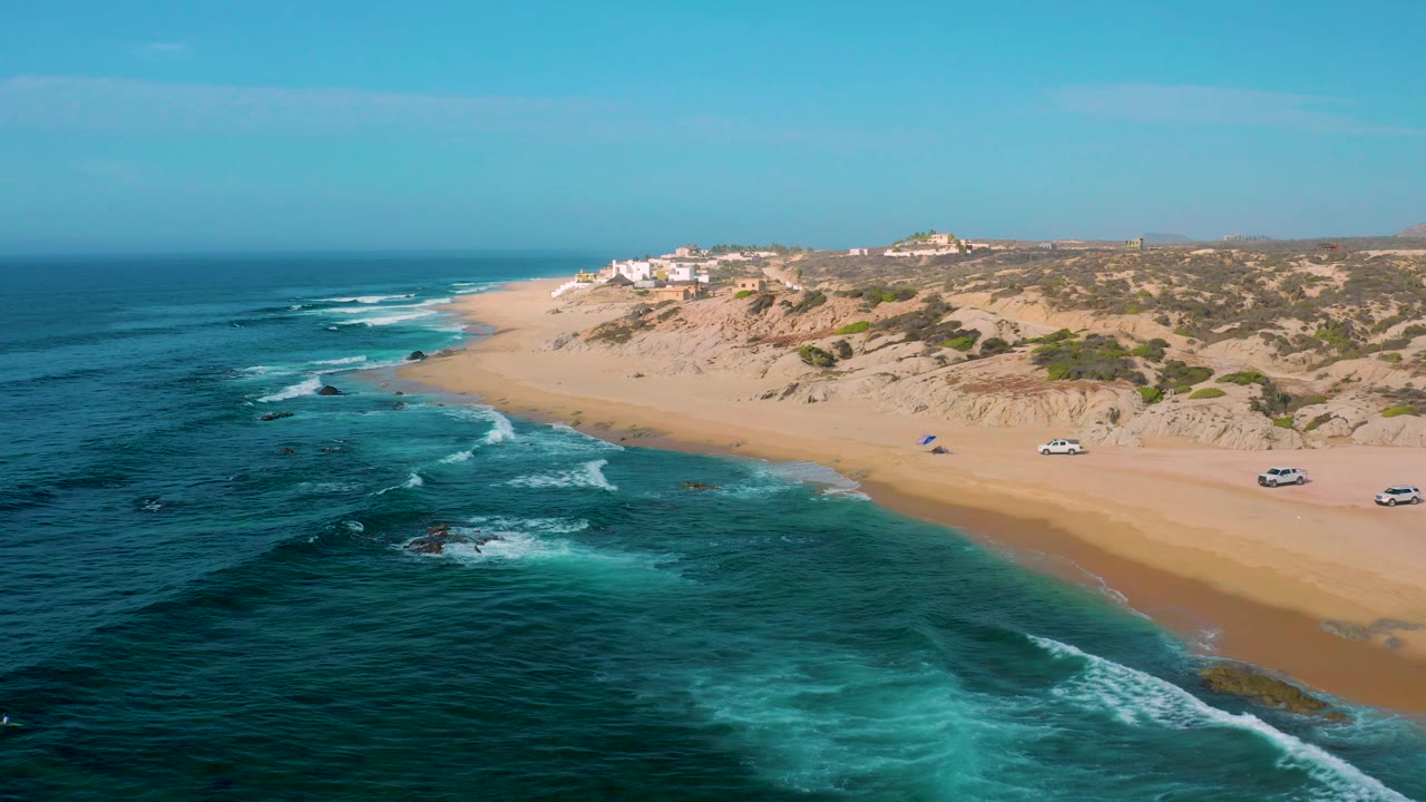 Aerial frame from Shipwrecks Beach with reef-darkened water in the foreground, white waves near shore, open sand, dunes, several small vehicles, and no visible people.