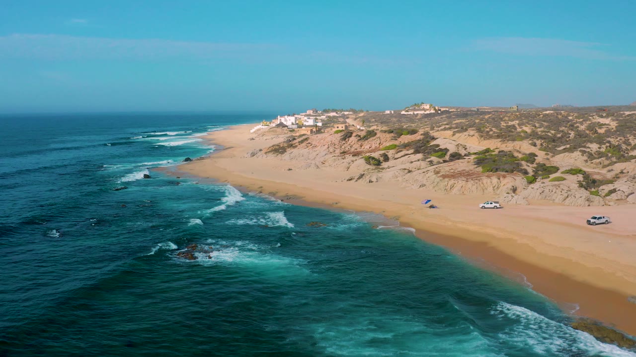 Aerial view of Shipwrecks Beach with the coastline running diagonally across frame, white surf in the lower left, pale sand to the right, scrubby dunes beyond, and no visible people.