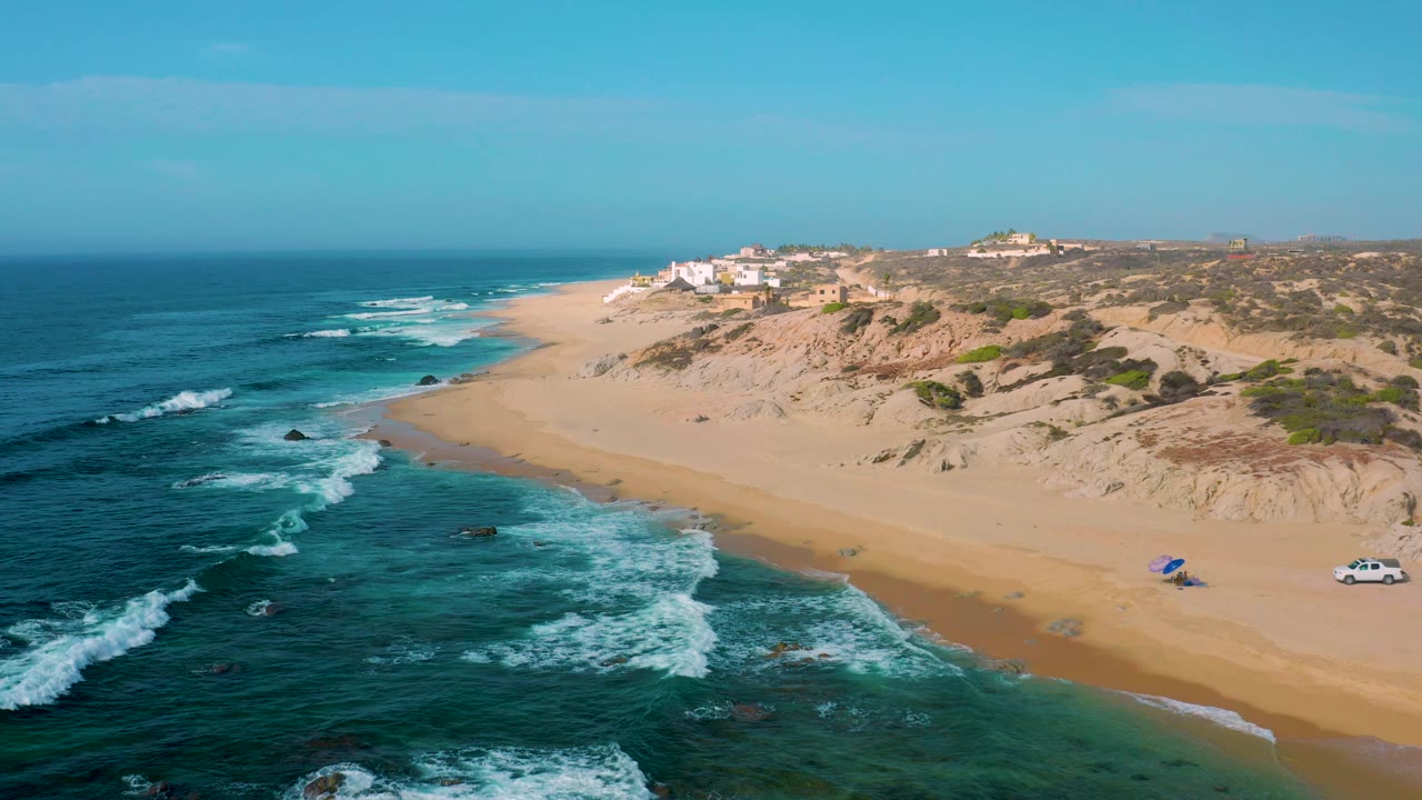 Aerial view at Shipwrecks Beach showing foamy waves pushing toward wet sand, dune slopes on the right, a small white vehicle near the beach edge, distant buildings, and no visible people.