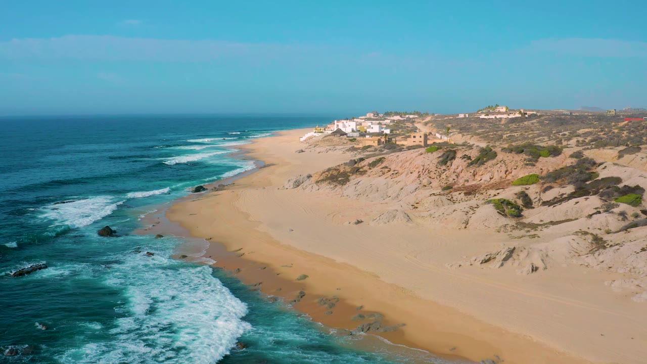 Shipwrecks Beach aerial frame showing a longer sweep of white breaking waves, pale sand curving toward dune slopes, small ridge buildings, blue sky, and no visible people.