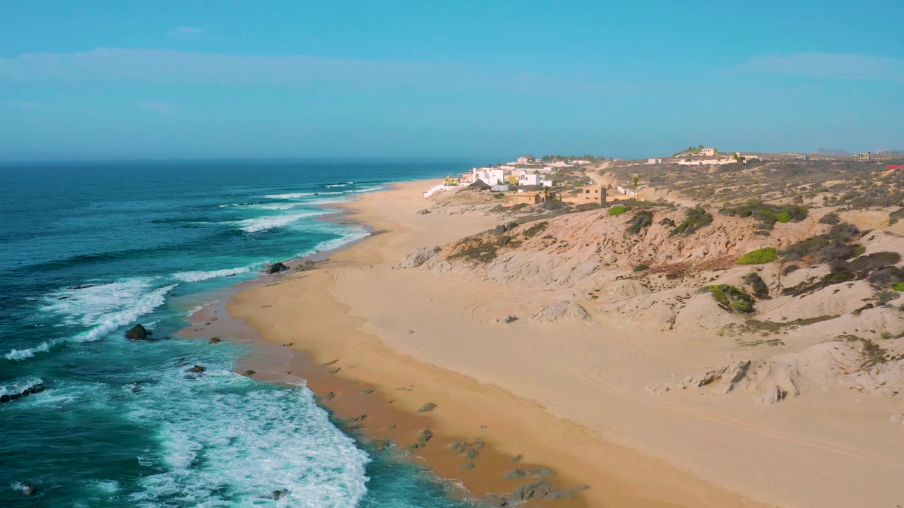 Aerial view over Shipwrecks Beach with foamy waves near dark rocks, an open sandy strip, tan dunes to the right, distant white structures on the ridge, and no visible people.