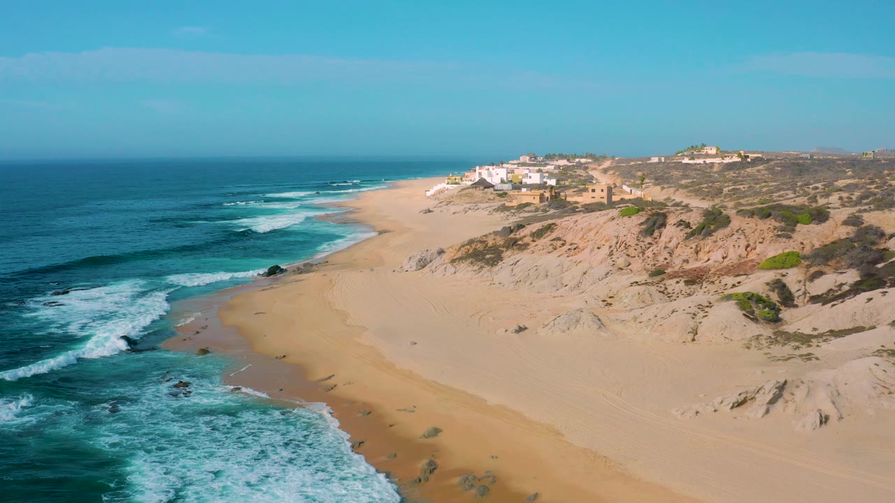 Shipwrecks Beach coastline from above, with ocean foam filling the lower left, a broad sandy beach through the center, scrub-covered dunes on the right, ridge buildings above, and no visible people.