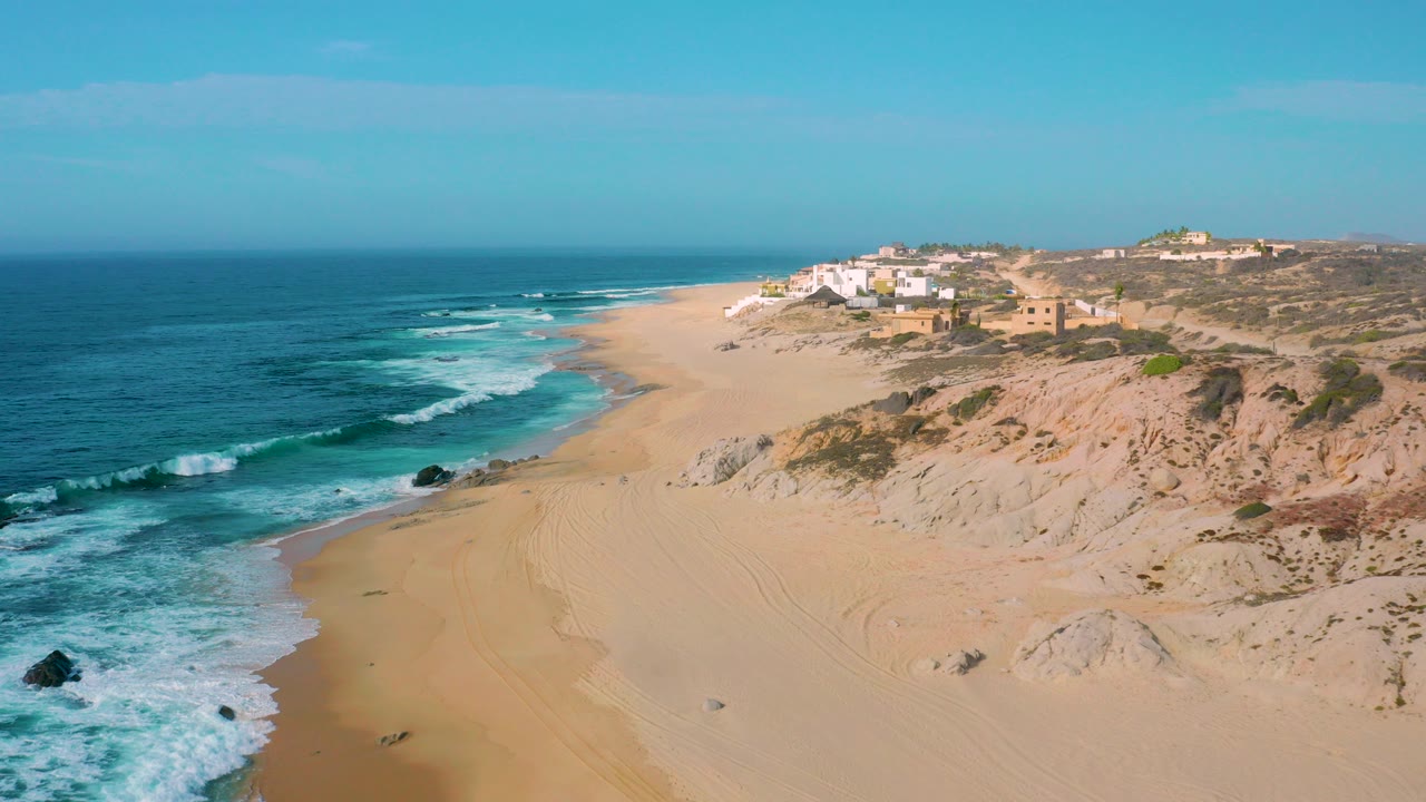 Aerial frame over Shipwrecks Beach in Baja California Sur with blue ocean on the left, pale sand and dunes on the right, bluff buildings in the distance, and no visible people.