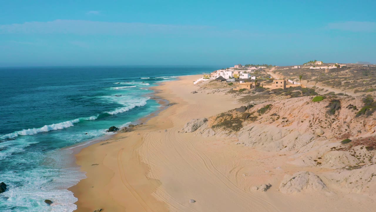 High view of Shipwrecks Beach with turquoise water, white wave bands near the shore, a mostly empty sandy beach, dune ridges, distant bluff buildings, and no visible people.