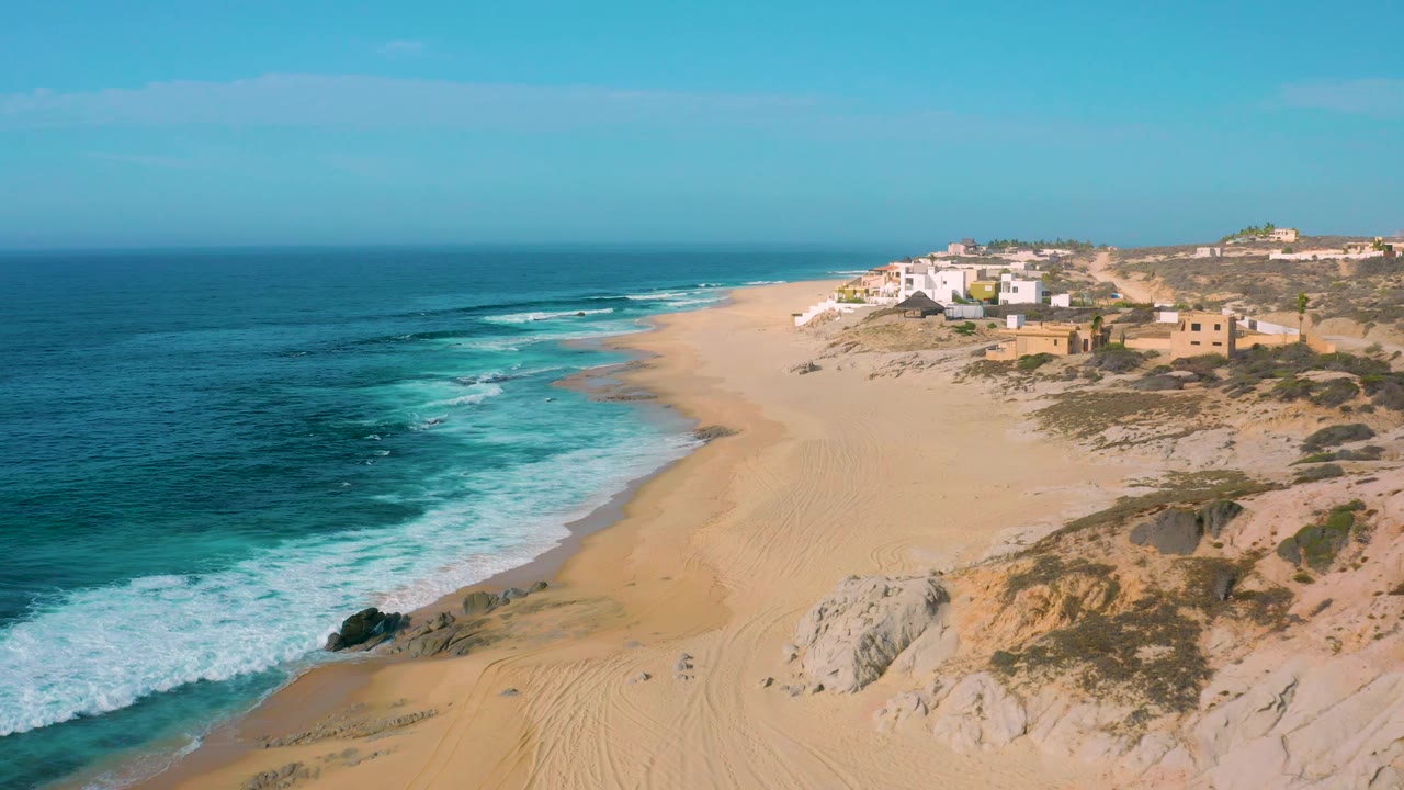 At Shipwrecks Beach, foamy blue waves push closer to the sand while pale rocks and low buildings remain visible along the right side, with no visible people.