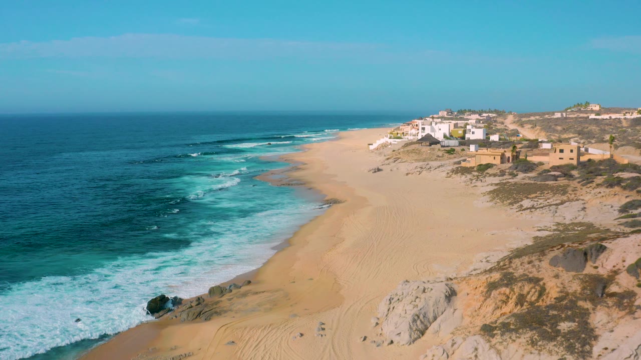 Wide aerial view of surf washing onto sand with rocky dunes in the foreground.