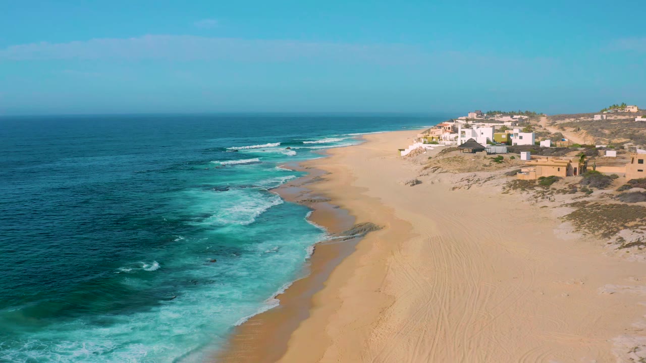 Shipwrecks Beach aerial frame with turquoise foam spreading along the left shoreline, broad tan sand to the right, low coastal homes on the bluff area, and no visible people.