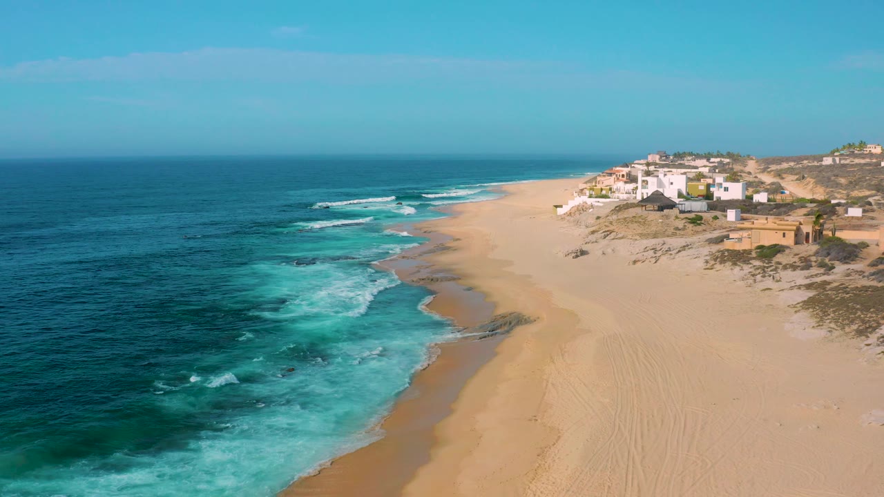 Aerial view at Shipwrecks Beach with darker offshore water, white foam reaching the sand, low dune vegetation near white buildings, and no visible people.