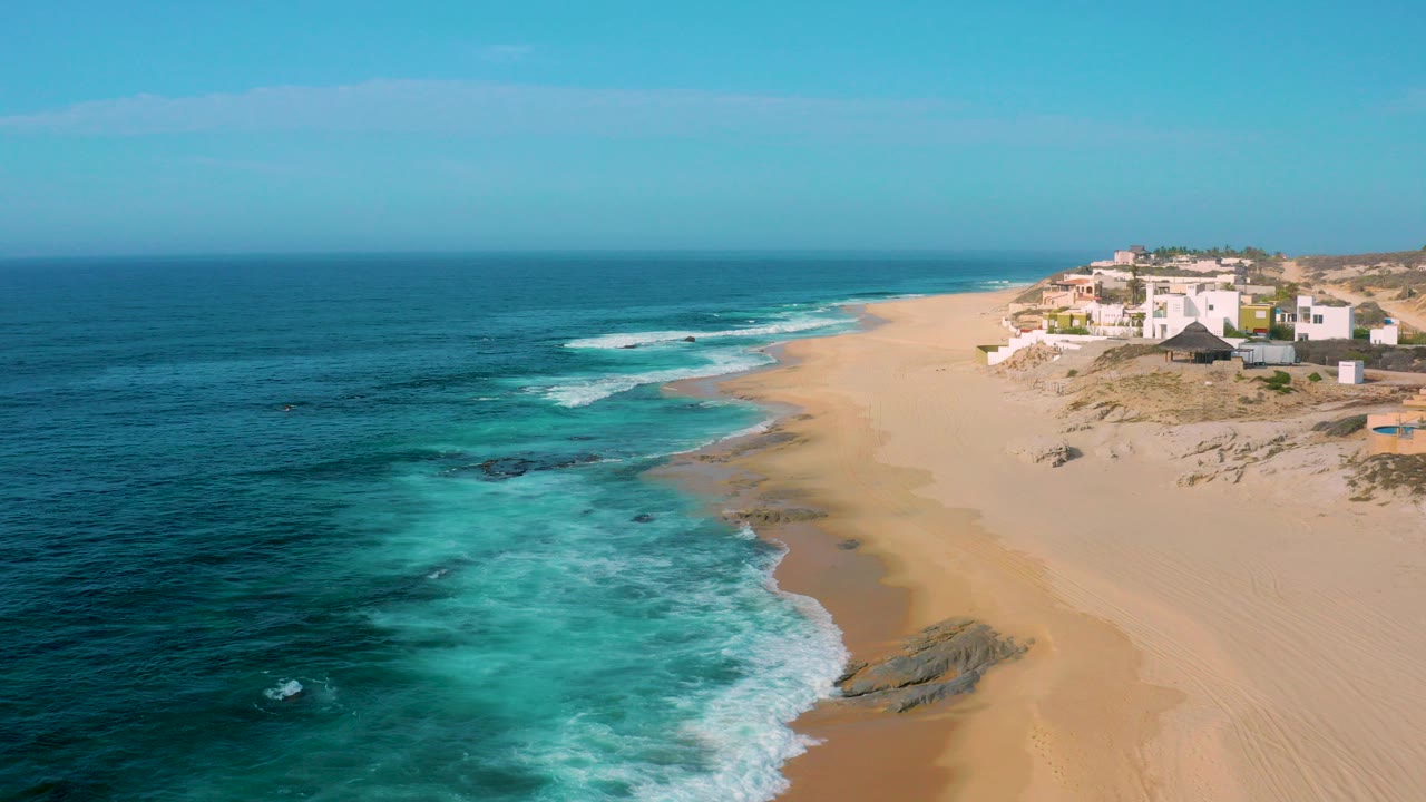 Aerial frame over Shipwrecks Beach, Baja California Sur, with turquoise surf on the left, tan sand curving toward white dune homes on the right, and no visible people.