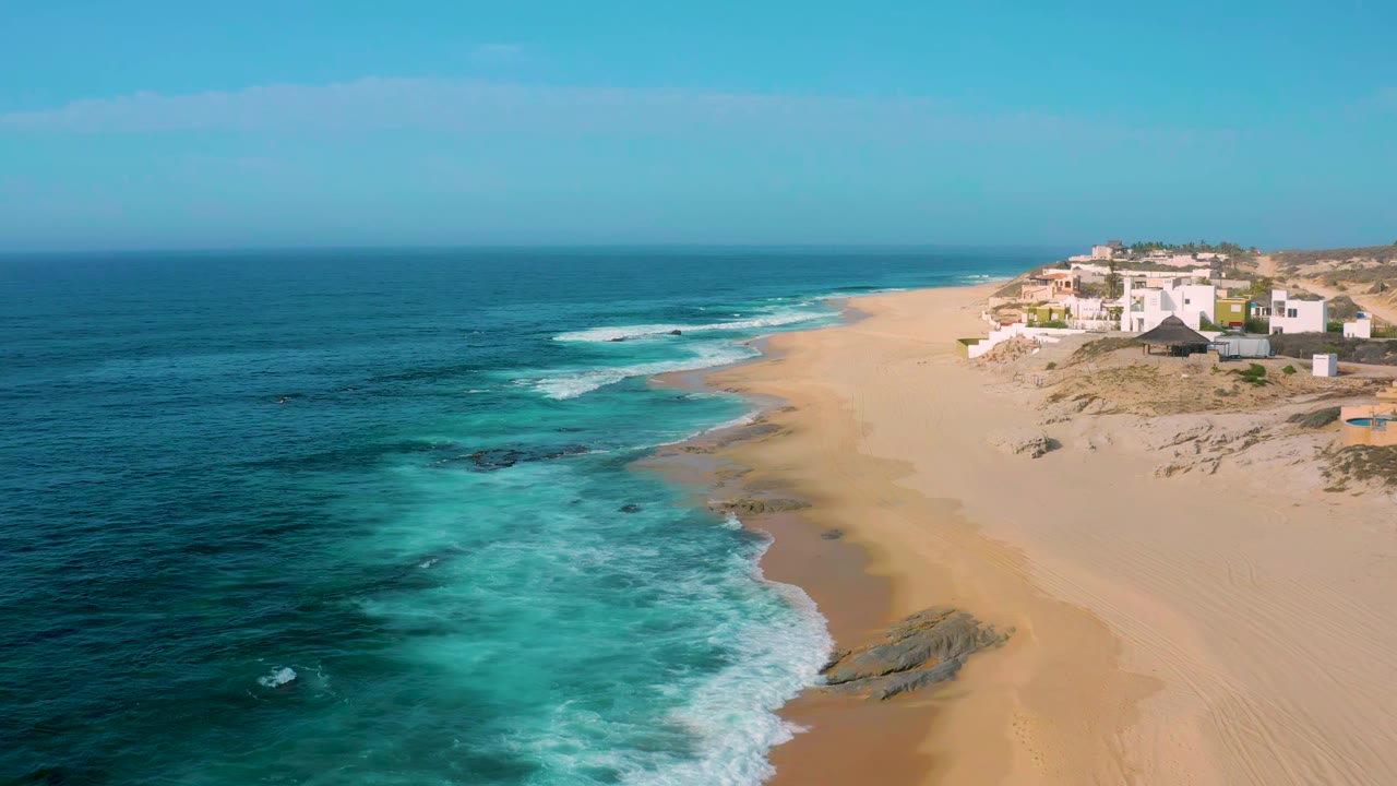 Aerial view at 0:01 showing foamy water spreading across the left foreground, a broad empty beach through the center, and white homes set back on the dune line at Shipwrecks Beach with no visible people.