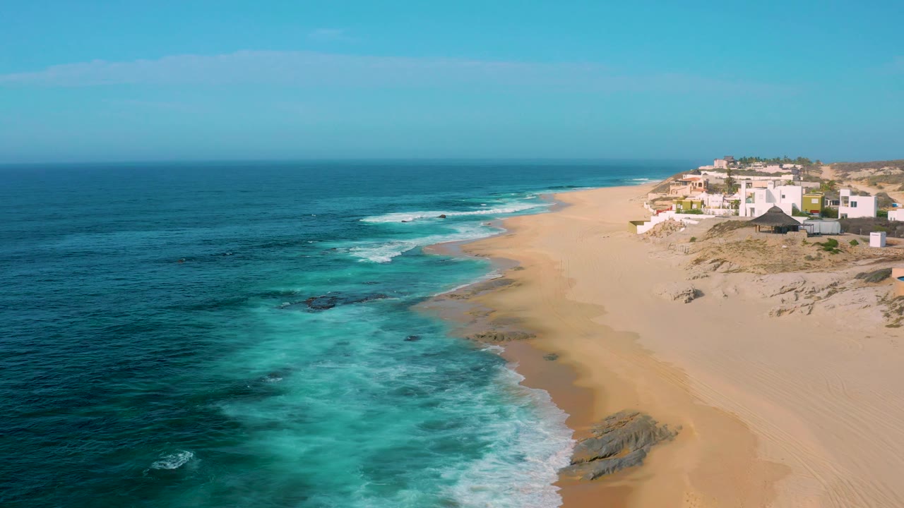 At 0:02, the aerial composition keeps the ocean across the left half, surf breaking near the center rocks, and a cluster of white dune homes at the upper right of Shipwrecks Beach, with no visible people.