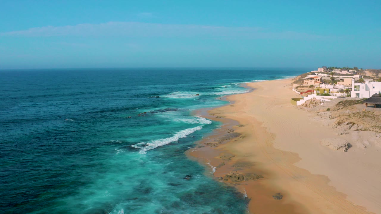Aerial view over Shipwrecks Beach in Baja California Sur, Mexico, with turquoise water at left, a broad sandy shoreline at center, coastal buildings on the right, and no visible people.