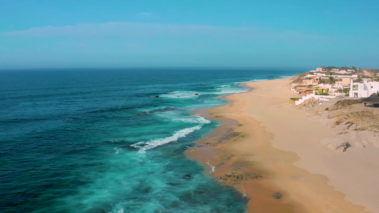 Wide aerial frame of Shipwrecks Beach with white surf bands reaching pale sand, darker rocks in the water, a cluster of coastal buildings at right, and no visible people.