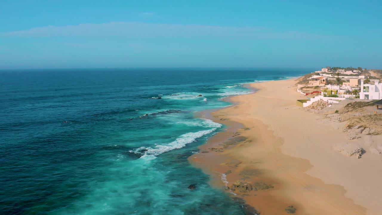Aerial view at Shipwrecks Beach showing deep blue ocean, bright turquoise shallows, a smooth sandy beach, white buildings on the right bluff area, and no visible people.