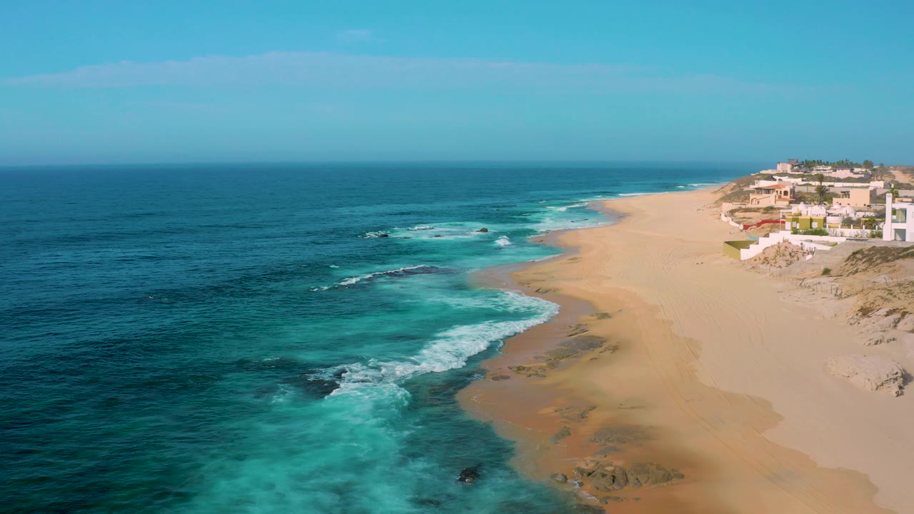 Aerial coastline view with turquoise water, rolling surf, sand, and buildings to the right.