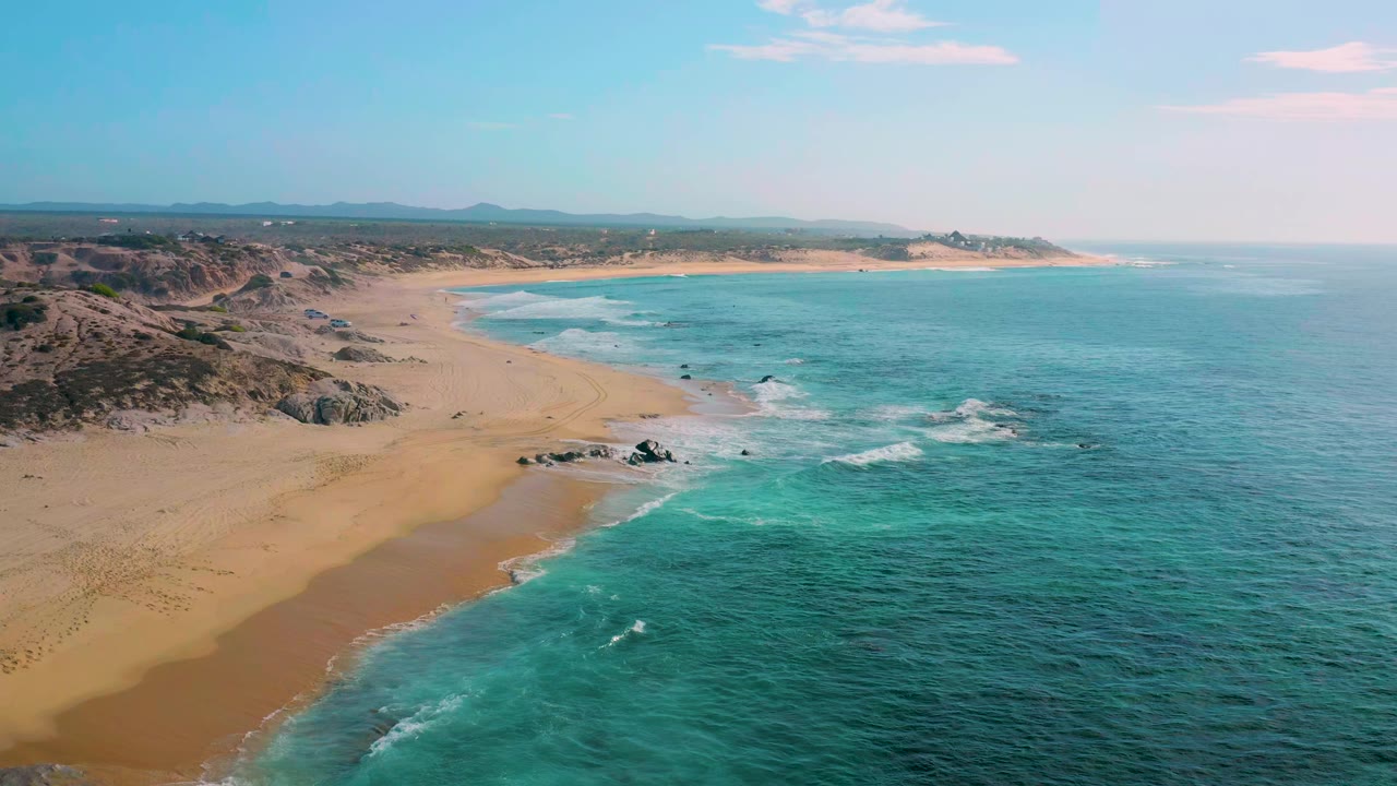 At Shipwrecks Beach in Baja California Sur, an aerial frame looks along a broad sand beach with blue-green surf in the foreground, rocky patches near shore, low dunes inland, and no visible people.