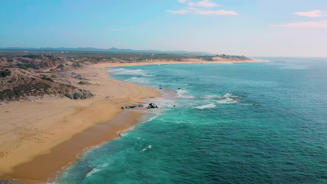 The second frame keeps the Shipwrecks Beach shoreline in a wide aerial view, with wave bands wrapping around dark rocks, tan beach stretching left, distant coastline ahead, and no visible people.