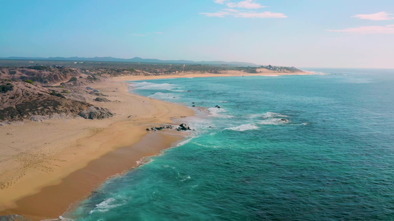 At 0:02, the aerial composition shows more open ocean on the right, a long tan shoreline on the left, small white breakers near exposed rocks, distant hills, and no visible people.
