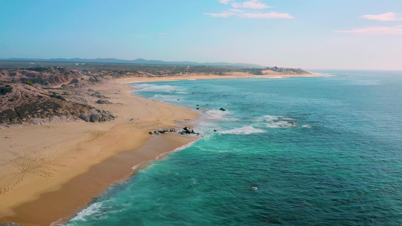 Midway through the clip, the frame looks across Shipwrecks Beach with a curved bay ahead, foamy surf tracing the sand, rocky outcrops in the nearshore, inland scrub, and no visible people.