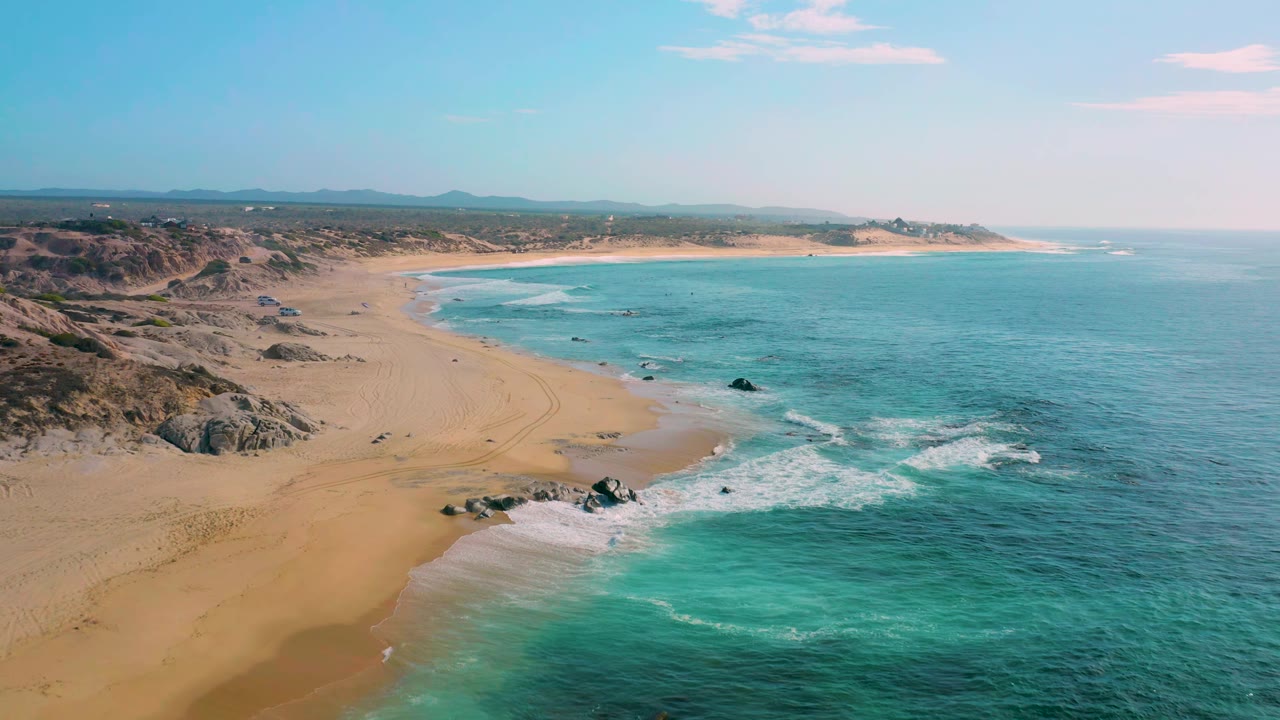 High aerial frame of Shipwrecks Beach, Baja California Sur, showing foamy surf reaching the sand, dark rocks in the shallows, low dunes inland, and no visible people.
