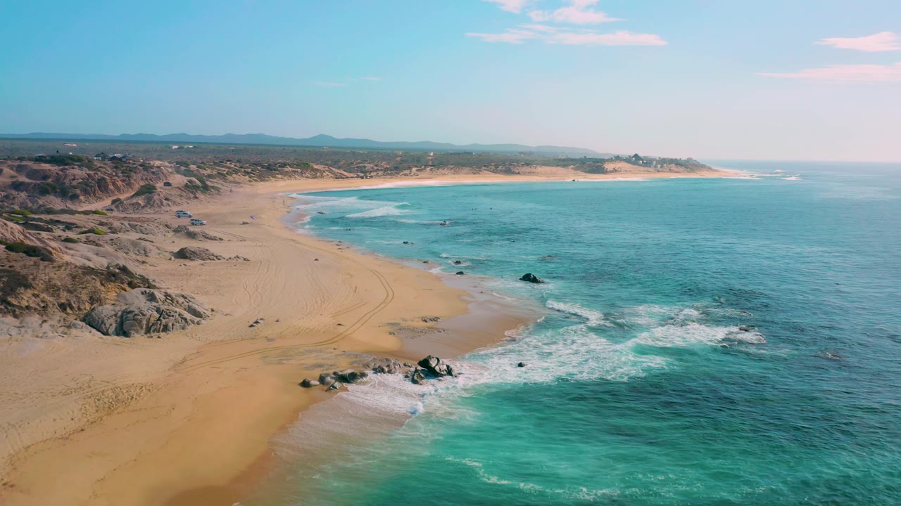 Aerial view of Shipwrecks Beach in Baja California Sur, with the shoreline curving through the frame, scattered rocks near the surf, broad empty sand, and no visible people.