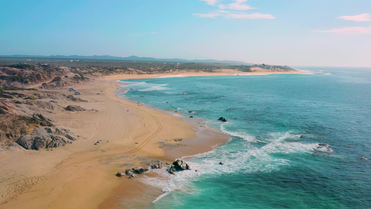 Wide aerial frame over Shipwrecks Beach, Baja California Sur, showing tire-like tracks in the sand, rocky dune formations to the left, surf on the right, and no visible people.