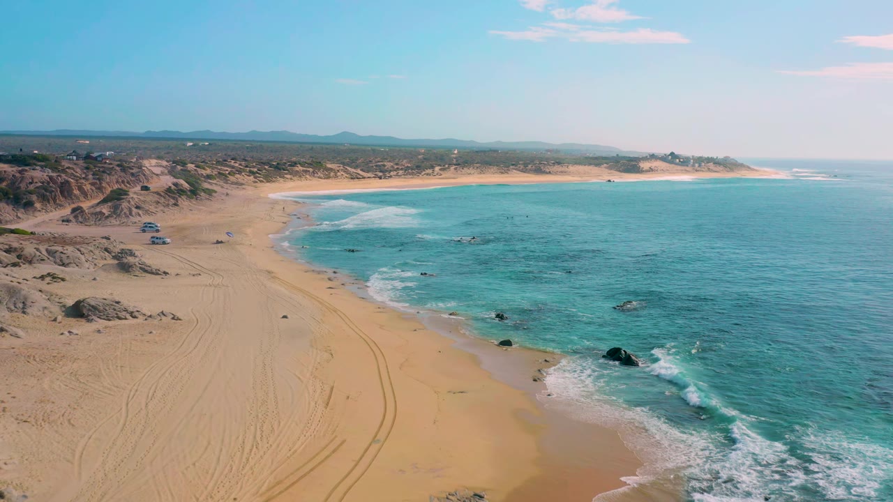 Wide drone view of curving beach, shallow water, and scattered rocks