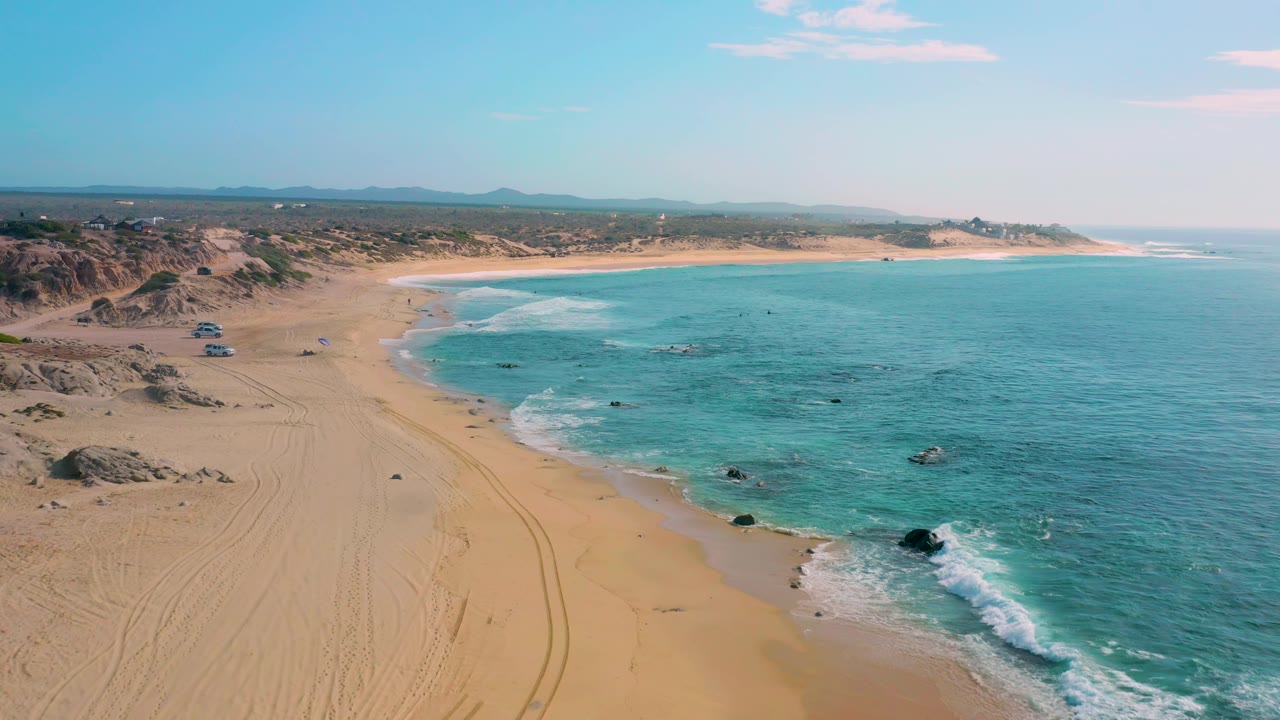 Aerial view over Shipwrecks Beach showing low surf along the sand, more open turquoise ocean to the right, dark rocks near the waterline, distant dunes, and no visible people.