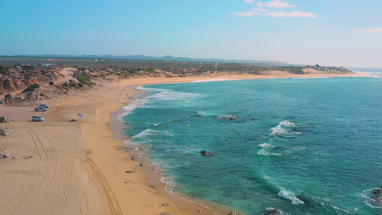 Aerial view over Shipwrecks Beach in Baja California Sur, with sandy shoreline, turquoise water, white surf, dune-backed terrain, parked vehicles, and tiny people visible near the water.