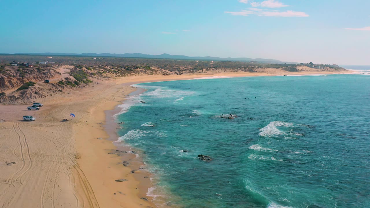 Aerial frame at Shipwrecks Beach with sand on the left, turquoise ocean on the right, foam lines crossing shallow water, exposed rocks, low dunes, parked vehicles, and small figures visible.