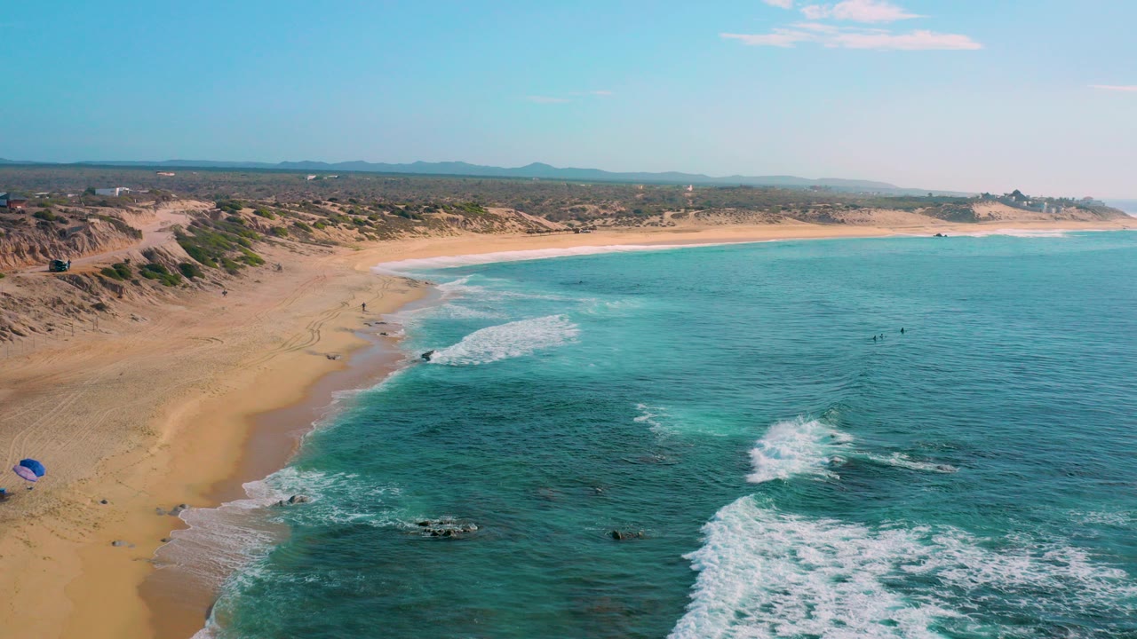 Aerial view over Shipwrecks Beach in Baja California Sur, with broad sand at left, turquoise water at right, white surf over dark rocks, distant dunes, and tiny people visible near the shoreline.