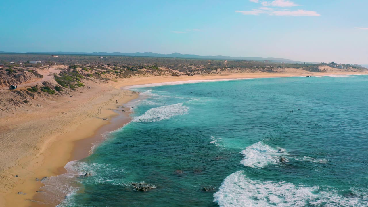 Drone-like aerial frame at Shipwrecks Beach with open turquoise water filling the right side, pale beach and dunes on the left, white surf breaking over rocks, and small people visible offshore.