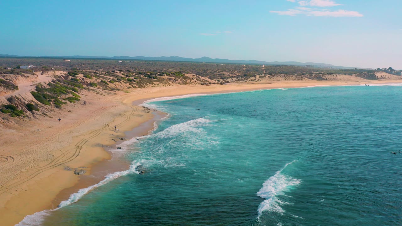 Aerial view over Shipwrecks Beach in Baja California Sur, with turquoise water washing onto a sandy shore, low scrub-covered dunes inland, distant hills, and no visible people.
