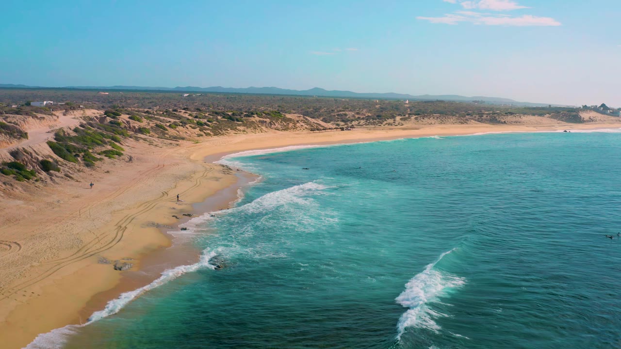 Shipwrecks Beach shoreline curves across the frame as clear blue water reaches the sand, with desert scrub behind the beach, a level horizon, and no visible people.