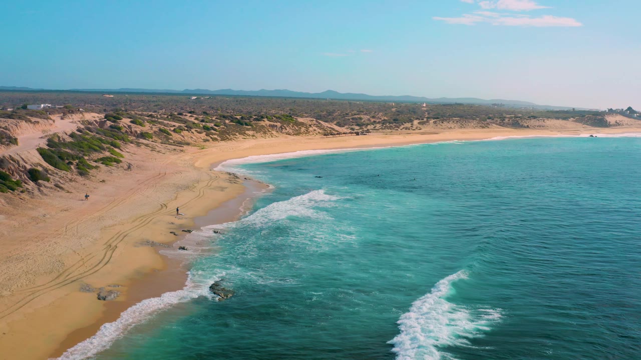 A wide drone-style view of Shipwrecks Beach shows turquoise water filling the right side, white wave bands near shore, sandy dunes to the left, distant hills, and no visible people.