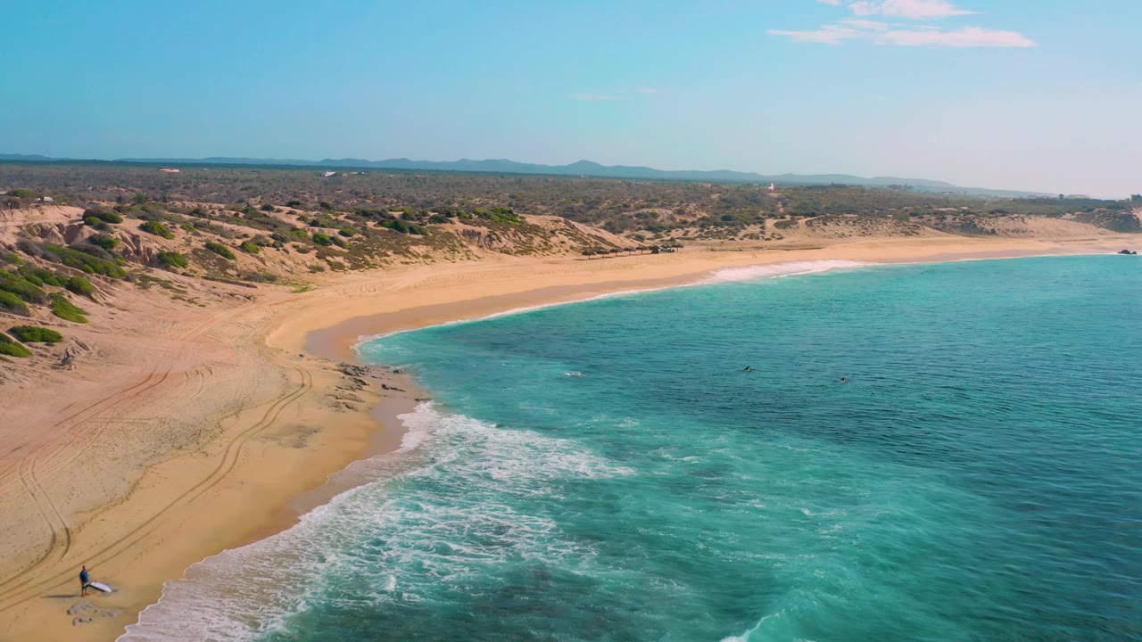 Shipwrecks Beach appears from above with turquoise water filling the right side, a pale sandy beach curving left, dune tracks in the sand, scrubland beyond, and tiny people visible near the lower-left shore.