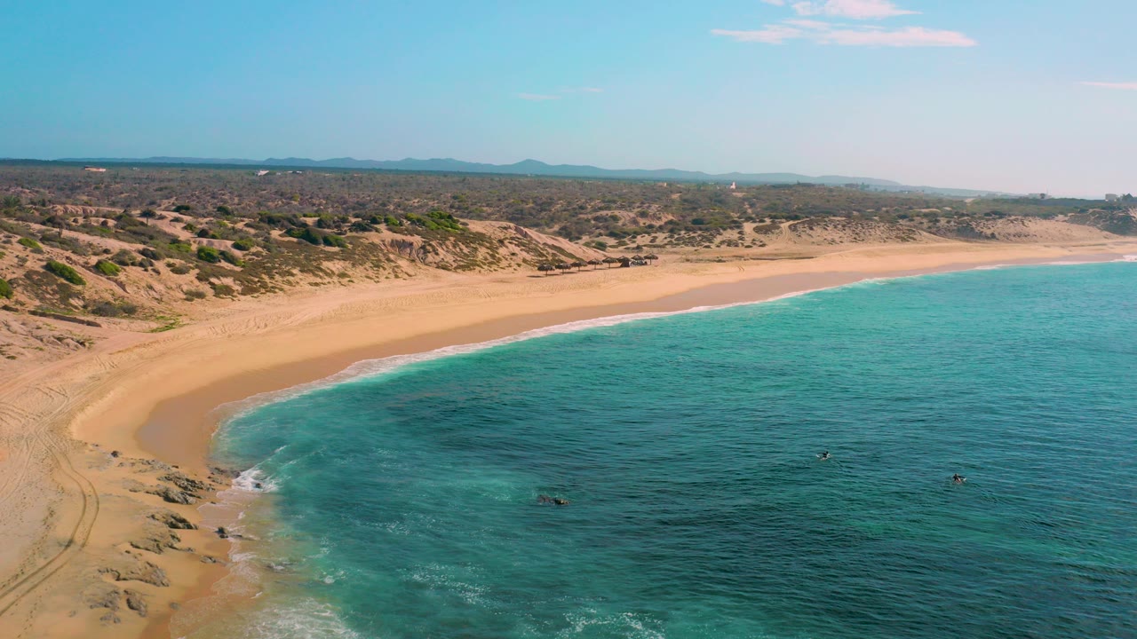 Aerial frame over Shipwrecks Beach in Baja California Sur showing a curved empty sandy shoreline, clear blue-green water, scrub dunes, distant hills, and no visible people.