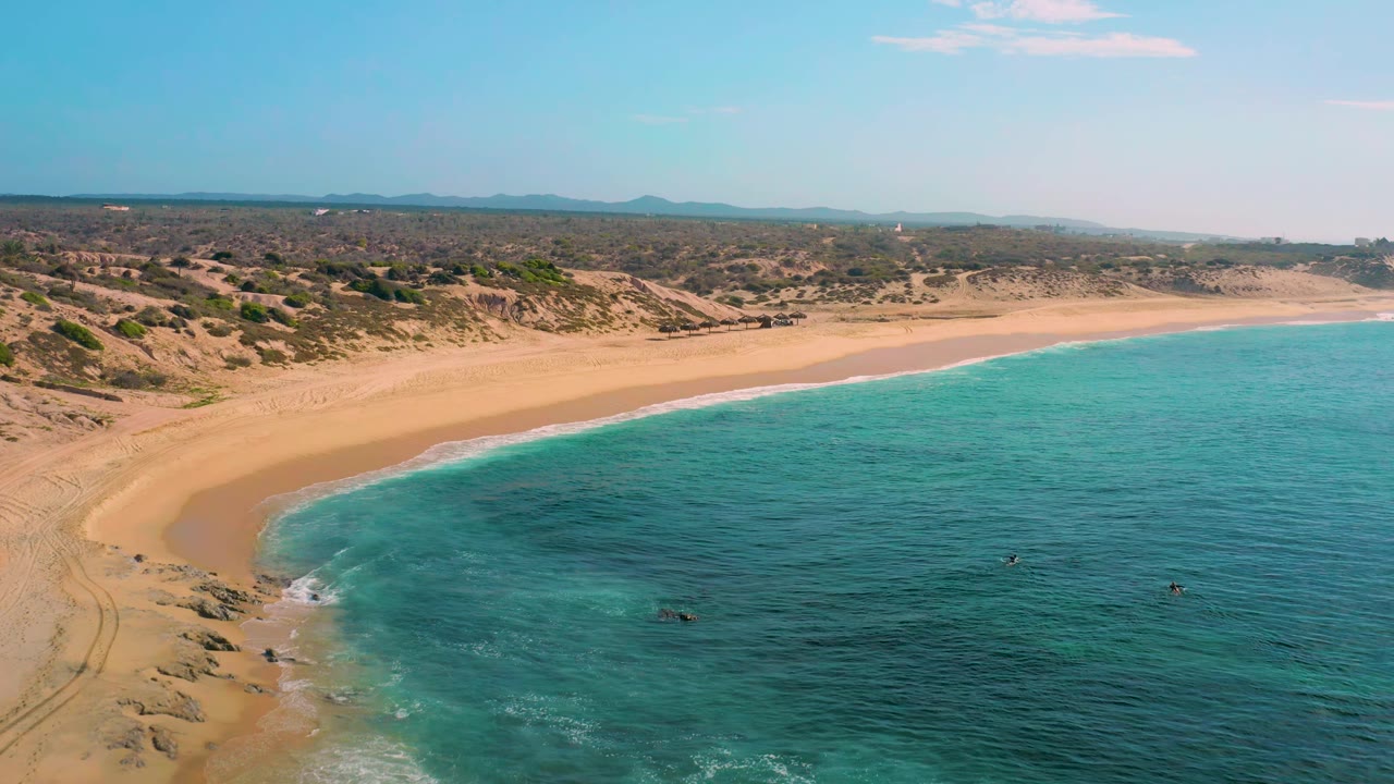 Aerial view at Shipwrecks Beach with turquoise water filling the foreground, a vacant sand beach along the left side, scrubby dunes inland, and no visible people.