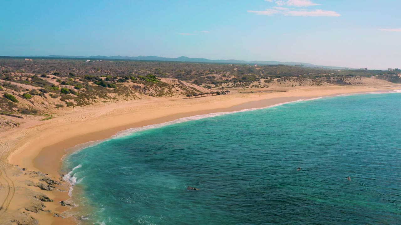 Wide aerial frame of Shipwrecks Beach showing the sandy coast sweeping across the image, calm blue-green sea to the right, dry inland terrain, distant hills, and no visible people.