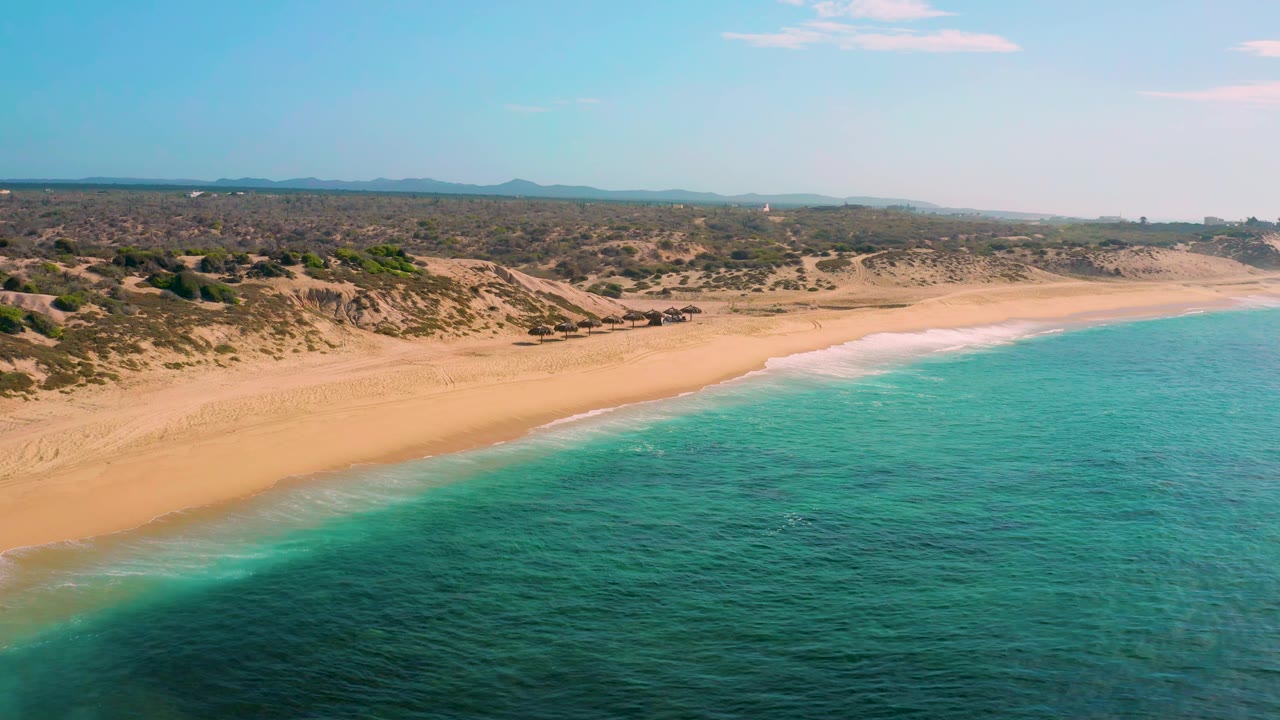 Aerial view at Shipwrecks Beach showing the shoreline angled across the frame, pale sand at low tide, dark patches under turquoise water, scrubby dunes inland, and no visible people.