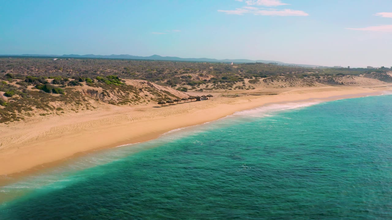 Shipwrecks Beach aerial frame with turquoise sea filling the lower right, a smooth band of empty sand, low vegetation on dunes, hazy hills on the horizon, and no visible people.