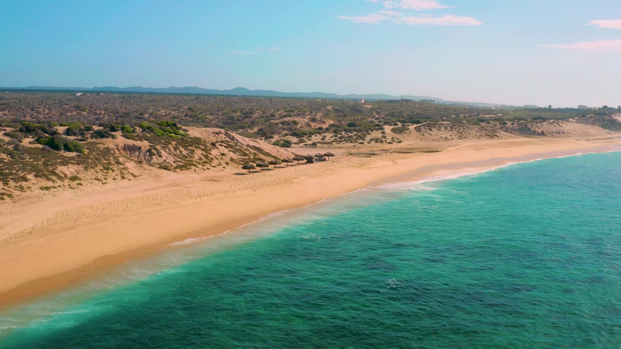 Mid-clip aerial over Shipwrecks Beach with the surf line running along the lower beach, turquoise water in the foreground, dry dunes and scrub inland, distant hills, and no visible people.